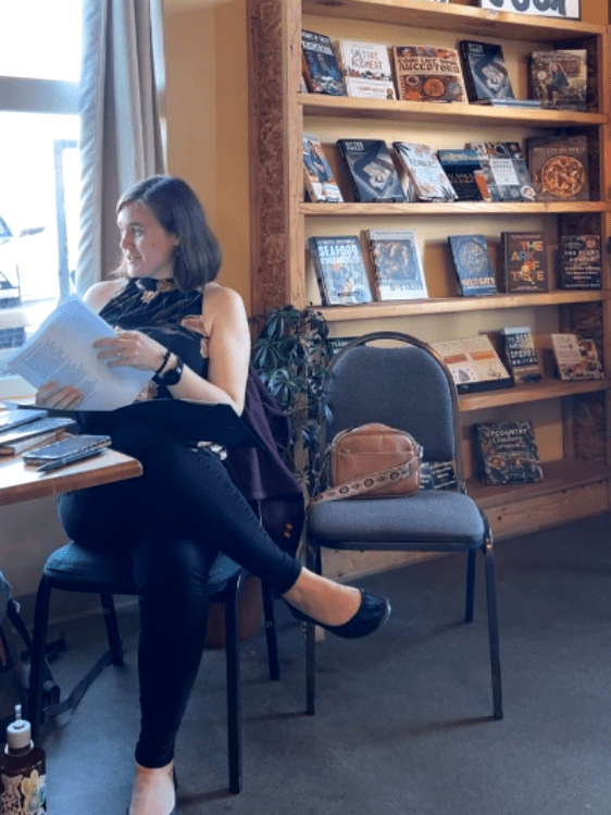 Caitlin holding papers sitting at a table in a chair by a window in front of a bookcase.